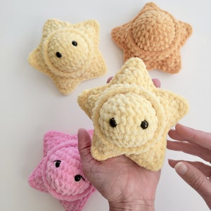 Puede incluir: Tres juguetes de peluche con forma de estrella hechos a crochet. Uno es amarillo, otro rosa y otro naranja. La estrella amarilla está siendo sostenida por una mano.