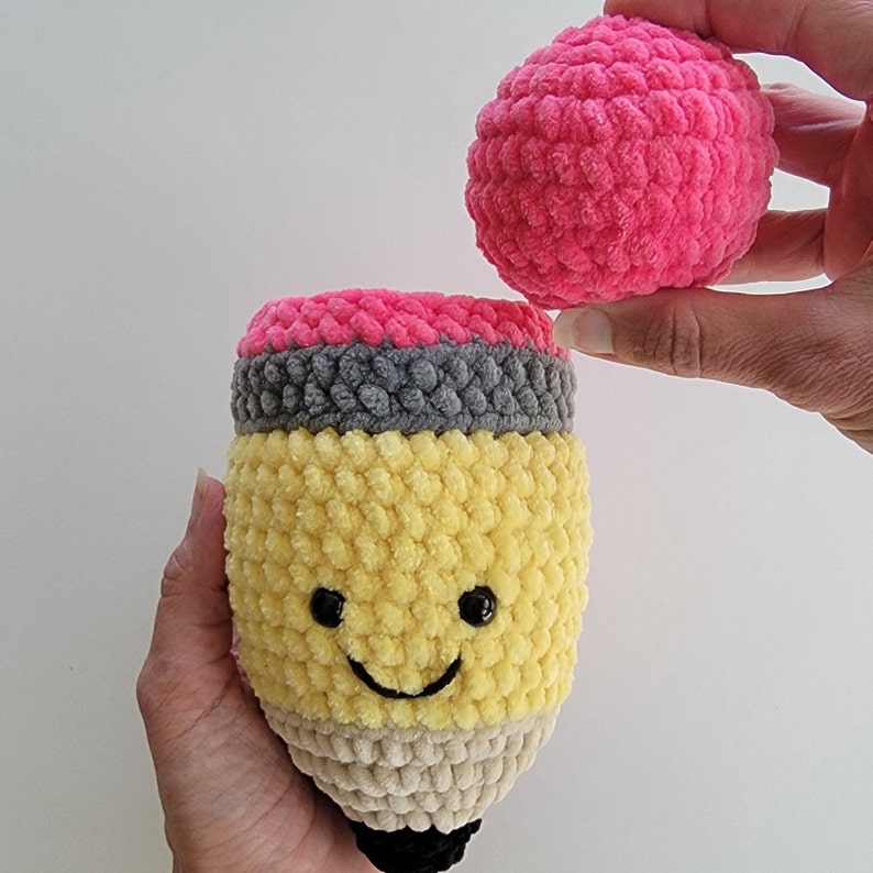 May include: A crocheted pencil toy with a yellow body, gray eraser, and pink top. The pencil has a smiling face and is holding a pink pom-pom.