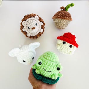 Può includere: Una collezione di cinque giocattoli all'uncinetto fatti a mano: un leone, una ghianda, un fungo, un agnello e una rana. Il leone ha una criniera marrone e una faccia bianca. La ghianda è marrone e beige. Il fungo ha un cappello rosso e bianco. L'agnello è bianco e la rana è verde.