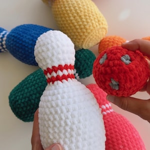May include: A set of crocheted bowling pins and a bowling ball made from soft, textured yarn. The pins are available in solid colours including red, yellow, blue, green, white, and pink, each featuring a contrasting band near the neck.