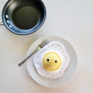 Puede incluir: Un huevo de crochet blanco con una yema de crochet amarilla y ojos negros. El huevo está en un plato blanco con un tenedor plateado.