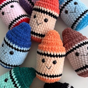 May include: A collection of plush, crocheted crayon-shaped toys in various colors including red, purple, peach, blue, orange, brown, teal, yellow, and black. Each soft, textured figure features two black bead eyes and a stitched black smile.