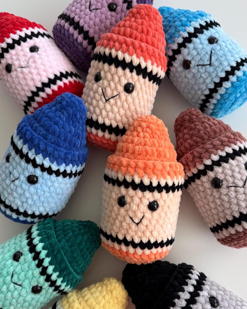 May include: A collection of plush, crocheted crayon-shaped toys in various colors including red, purple, peach, blue, orange, brown, teal, yellow, and black. Each soft, textured figure features two black bead eyes and a stitched black smile.