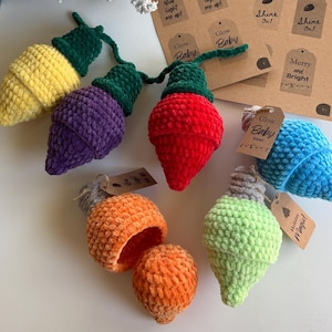 May include: A collection of colorful, crocheted Christmas light ornaments in yellow, purple, red, orange, blue, and green. Each light has a green or gray base and a loop for hanging. Some lights have small tags that say "Glow Baby" and "Holiday Magic!"
