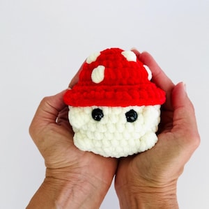 Puede incluir: Un juguete de hongo tejido a ganchillo con un gorro rojo con lunares blancos y un tallo blanco. El hongo tiene dos ojos negros. El juguete se sostiene en dos manos sobre un fondo blanco.