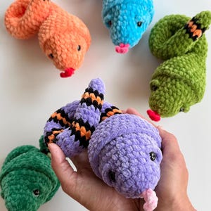 May include: A collection of five plush snakes made from soft, textured yarn in vibrant colours including orange, blue, green, and purple. Each snake features small dark eyes and a protruding yarn tongue. One purple snake is held in the foreground.