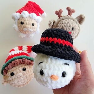 May include: Four crocheted plush ornaments featuring festive characters: a Santa Claus with a red hat, a reindeer with brown antlers, an elf with a red and white striped hat, and a snowman wearing a black hat with a red band.