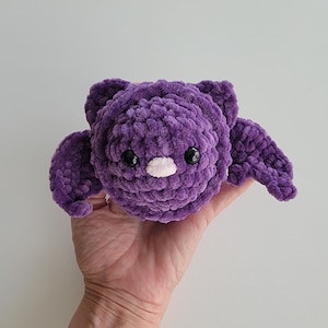 May include: A purple crocheted bat plush toy with black eyes and a pink nose. The toy is being held in a person's hand.