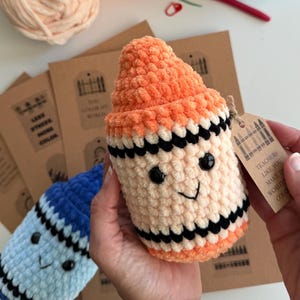May include: A plush crochet crayon toy in orange and cream with black stripes, featuring black safety eyes and a stitched smile. A brown paper tag attached with twine reads: Teachers like you make the world colorful, with a crayon graphic.