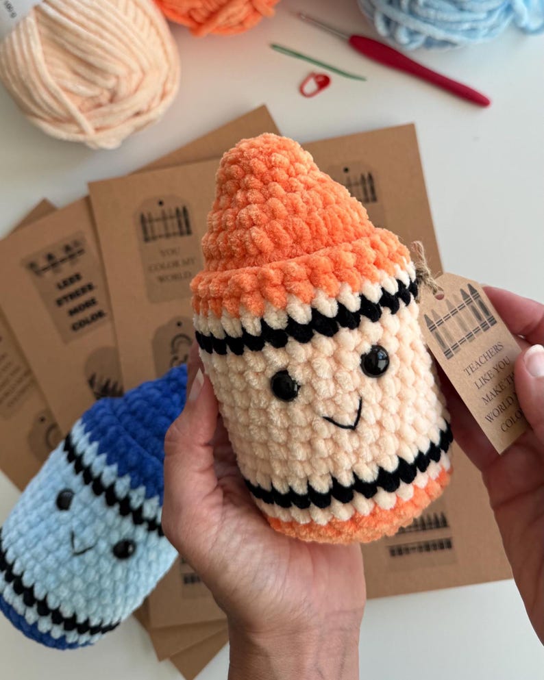 May include: A plush crochet crayon toy in orange and cream with black stripes, featuring black safety eyes and a stitched smile. A brown paper tag attached with twine reads: Teachers like you make the world colorful, with a crayon graphic.