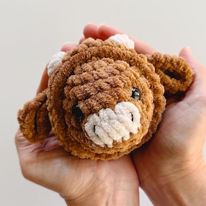 May include: A brown crocheted stuffed animal shaped like a cow with white yarn accents for the ears and nose. The cow has black eyes and a white nose.