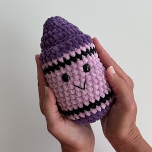 May include: A plush, crocheted crayon toy in shades of purple and light lavender. The crayon features a pointed top, two black plastic bead eyes, and a stitched black mouth. It is decorated with horizontal black zigzag stripes near the top and bottom.