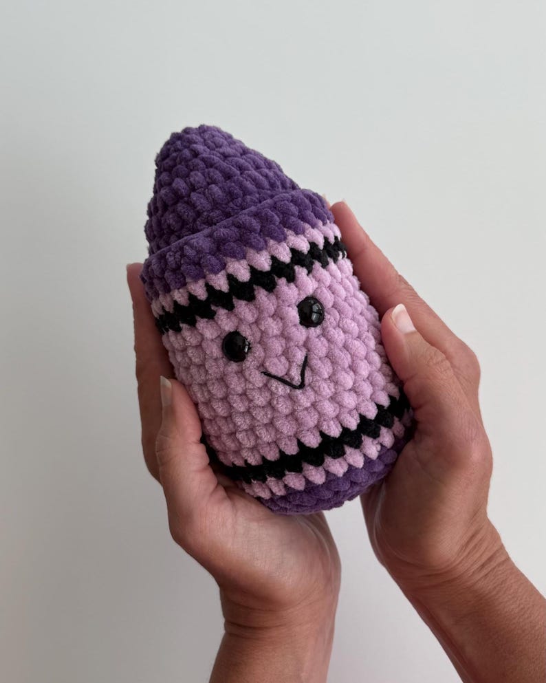 May include: A plush, crocheted crayon toy in shades of purple and light lavender. The crayon features a pointed top, two black plastic bead eyes, and a stitched black mouth. It is decorated with horizontal black zigzag stripes near the top and bottom.