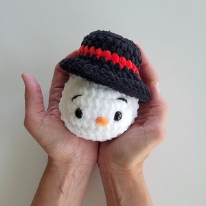 May include: A round, white crocheted plush snowman head featuring a black top hat with a red band. The snowman has two black plastic safety eyes, embroidered black eyebrows, and a small, orange crocheted carrot nose centred on the face.