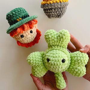 Könnte beinhalten: Drei gehäkelte Plüschtiere: ein grünes Kleeblatt, ein Kobold mit grünem Hut und orangem Haar und ein grauer Topf mit gelbem Rand.