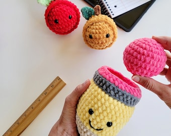 May include: A set of crocheted plush items including a red apple, a tan orange, and a yellow pencil-shaped container with a pink eraser top. A wooden ruler is visible, showing the pencil container is approximately 5 inches in height.