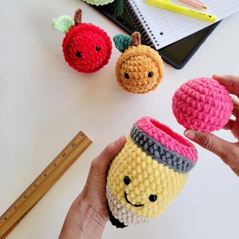 May include: A set of crocheted plush toys shaped like an apple, a pencil, and an orange. The pencil is open and has a pink crocheted lid. The toys are made with yarn in shades of red, yellow, orange, and pink.