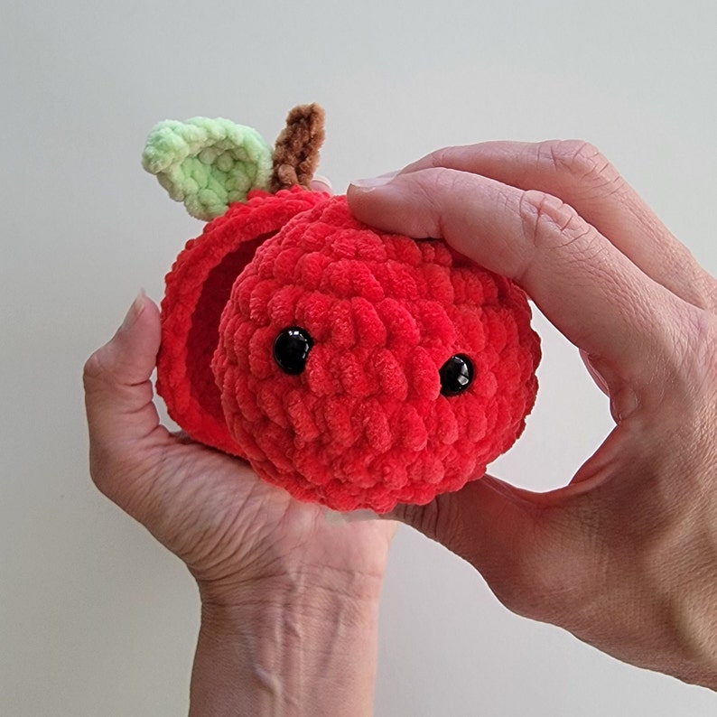 May include: A red crocheted apple with a green leaf and black eyes. The apple is open and appears to be a small box.