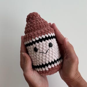 May include: A plush crochet toy shaped like a crayon, featuring a dusty rose top and base, a cream-colored center, and two horizontal black stripes. The toy has two small black plastic eyes and a stitched black V-shaped mouth on the front.
