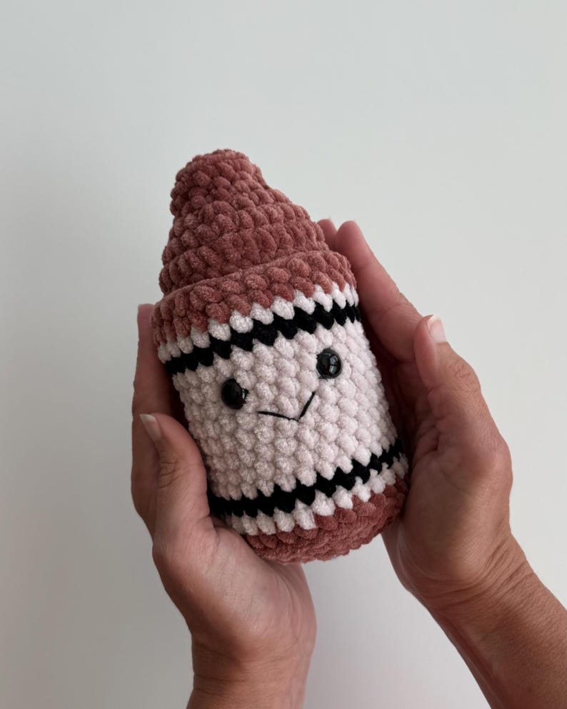 May include: A plush crochet toy shaped like a crayon, featuring a dusty rose top and base, a cream-colored center, and two horizontal black stripes. The toy has two small black plastic eyes and a stitched black V-shaped mouth on the front.