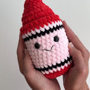 May include: A plush crochet toy shaped like a crayon with a red pointed tip, a light pink cylindrical body, and black horizontal stripes. The toy features two small black plastic eyes and a stitched black mouth, creating a simple facial expression.