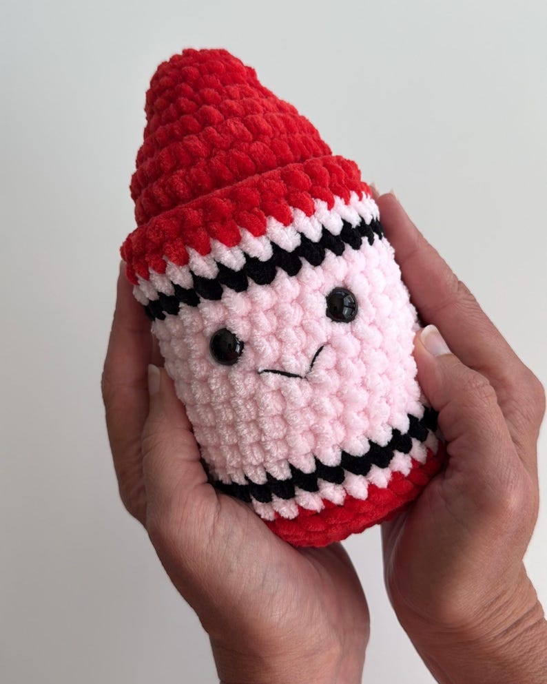 May include: A plush crochet toy shaped like a crayon with a red pointed tip, a light pink cylindrical body, and black horizontal stripes. The toy features two small black plastic eyes and a stitched black mouth, creating a simple facial expression.