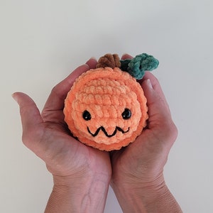 May include: A crocheted orange pumpkin with a green stem and a cute face. The pumpkin is being held in two hands.