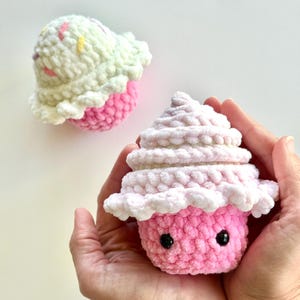 Puede incluir: Dos peluches de cupcake tejidos a crochet. Uno es rosa con glaseado blanco y el otro es blanco con glaseado rosa y chispas de colores. Ambos tienen ojos negros.