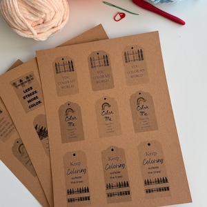 May include: A sheet of brown kraft paper tags featuring printed black graphics and text including "YOU COLOR MY WORLD," "Color Me," and "Keep Coloring outside the lines!" alongside yarn balls, a red crochet hook, and a green sewing needle.