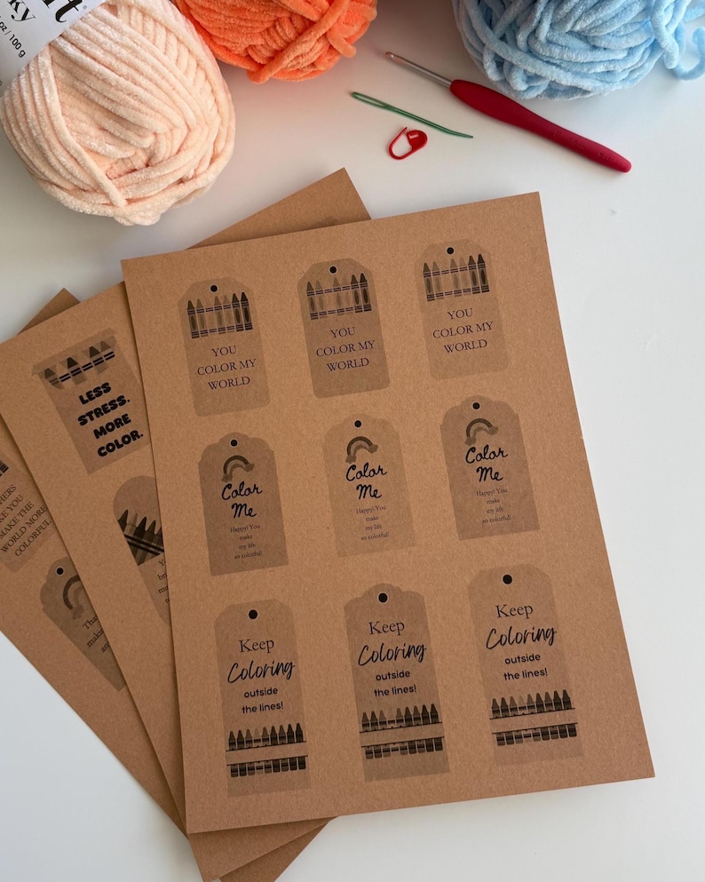 May include: A sheet of brown kraft paper tags featuring printed black graphics and text including "YOU COLOR MY WORLD," "Color Me," and "Keep Coloring outside the lines!" alongside yarn balls, a red crochet hook, and a green sewing needle.