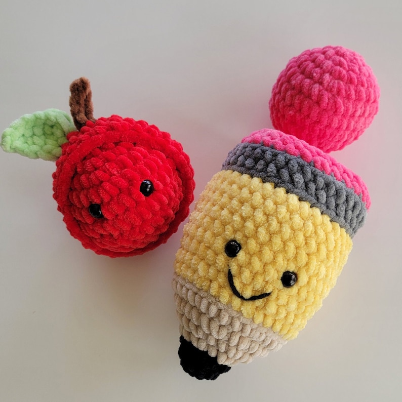 May include: A set of three crocheted plush toys: a red apple, a yellow pencil, and a pink eraser. The toys have smiling faces and are made with soft yarn.