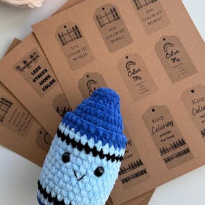May include: A blue and light blue crocheted plush crayon with black safety eyes and a stitched mouth, positioned over a stack of brown paper gift tags featuring black printed crayon graphics and phrases like You Color My World and Color Me.