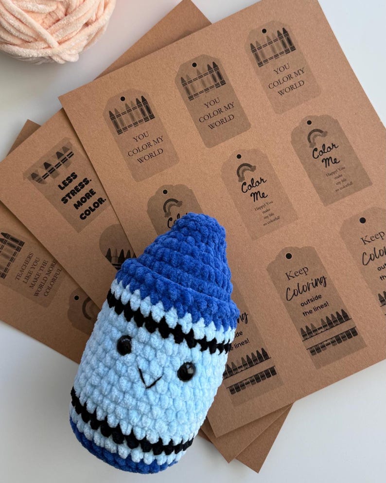May include: A blue and light blue crocheted plush crayon with black safety eyes and a stitched mouth, positioned over a stack of brown paper gift tags featuring black printed crayon graphics and phrases like You Color My World and Color Me.