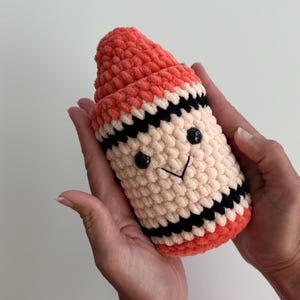 May include: A plush, crocheted toy shaped like a crayon, featuring a coral-colored top and base, a cream-colored middle section, and black horizontal stripes. The toy has two small, round black plastic eyes and a stitched black V-shaped mouth.