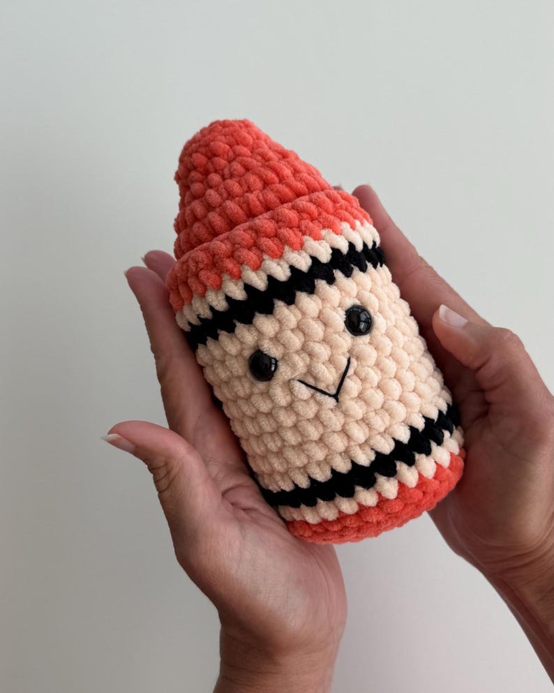 May include: A plush, crocheted toy shaped like a crayon, featuring a coral-colored top and base, a cream-colored middle section, and black horizontal stripes. The toy has two small, round black plastic eyes and a stitched black V-shaped mouth.
