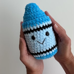 May include: A plush crochet crayon toy in shades of bright blue and light blue, featuring a pointed tip, black horizontal stripes, and a stitched face with two small black plastic eyes and a simple black embroidered smile on the light blue body.