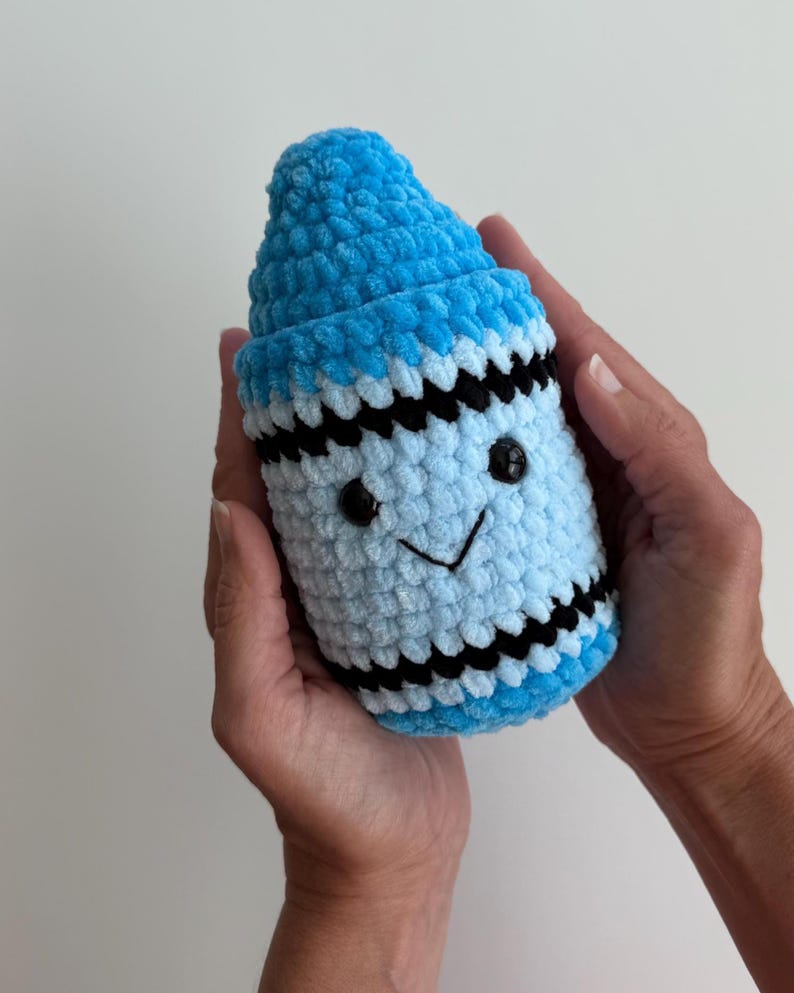 May include: A plush crochet crayon toy in shades of bright blue and light blue, featuring a pointed tip, black horizontal stripes, and a stitched face with two small black plastic eyes and a simple black embroidered smile on the light blue body.