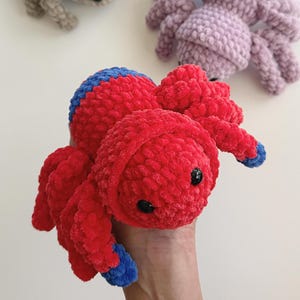 Puede incluir: Un juguete de araña de peluche hecho a mano en rojo vibrante con detalles azules en la cabeza y las patas. La araña tiene ojos negros de botón y un aspecto suave y texturizado. Otras arañas en gris y lavanda son visibles en el fondo.