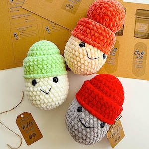 May include: Three crocheted pill-shaped plushies with smiling faces and hats. The plushies are in green, orange, and red. The plushies are sitting on a brown surface with a tag that says "I have pills for that!"