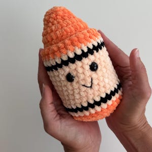 May include: A plush crochet toy shaped like a crayon, featuring a bright orange pointed tip, a cream-colored body, and black horizontal stripes. The toy has two small black plastic eyes and a stitched black smile on the front surface.
