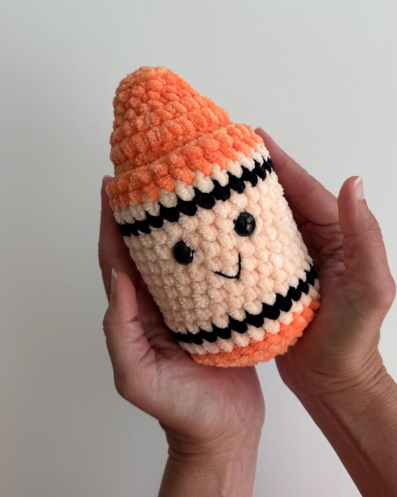 May include: A plush crochet toy shaped like a crayon, featuring a bright orange pointed tip, a cream-colored body, and black horizontal stripes. The toy has two small black plastic eyes and a stitched black smile on the front surface.