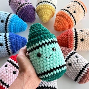 May include: A collection of plush, crocheted crayon-shaped toys in various colors including purple, yellow, orange, red, pink, blue, brown, and green. Each soft toy features a stitched black and white band, black plastic eyes, and a small smile.