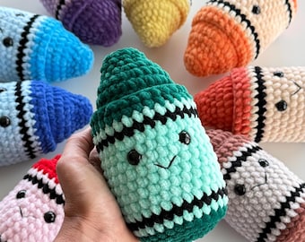 May include: A collection of plush, crocheted crayon-shaped toys in various colors including mint green, purple, yellow, orange, blue, pink, and brown. Each soft toy features a stitched smiling face with black eyes and a black embroidered mouth.