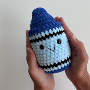 May include: A plush crochet toy shaped like a crayon, featuring a dark blue pointed tip, a light blue cylindrical body, and horizontal black stripes. The toy has two small black plastic eyes and a stitched black V-shaped mouth on the front.