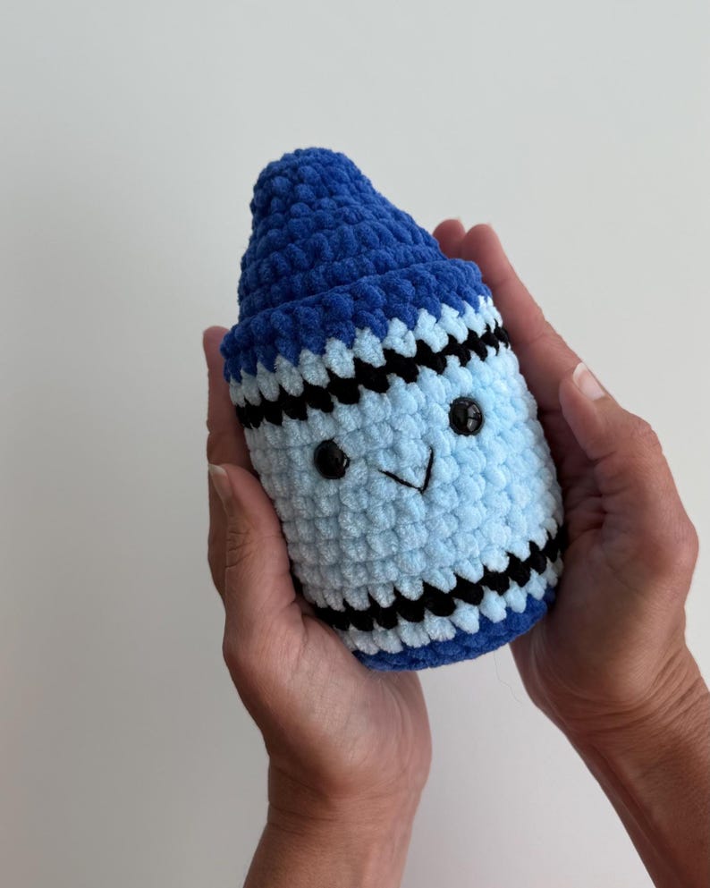 May include: A plush crochet toy shaped like a crayon, featuring a dark blue pointed tip, a light blue cylindrical body, and horizontal black stripes. The toy has two small black plastic eyes and a stitched black V-shaped mouth on the front.