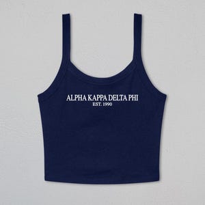 May include: A navy blue sleeveless crop top with a scoop neckline and thick shoulder straps. The center of the chest features white text that reads ALPHA KAPPA DELTA PHI and EST. 1990 in a clean, capitalized serif font style.