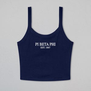 May include: A navy blue cropped tank top featuring a scoop neckline and thick shoulder straps. The center of the chest displays the white text "PI BETA PHI" in a serif font, with the smaller white text "EST. 1867" positioned directly below it.