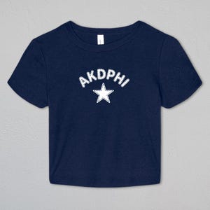 May include: A navy blue short-sleeved crop top featuring the text AKDPHI in white letters with a stitched-style outline, positioned above a white five-pointed star with a matching stitched-style outline centered on the chest area.
