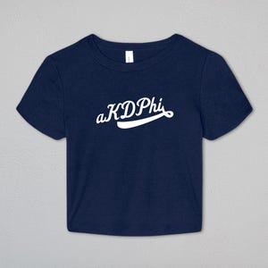 May include: A navy blue short-sleeved crop top featuring the white text aKDPhi printed in a cursive script font across the center of the chest. The garment has a crew neckline and a straight hemline, providing a simple and clean design.