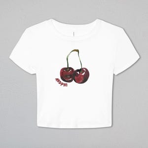 May include: A white short-sleeved crop top featuring a watercolor-style illustration of two red cherries with green stems. The text AKDPHI is printed in a dark red font underneath the cherries on the front of the garment.